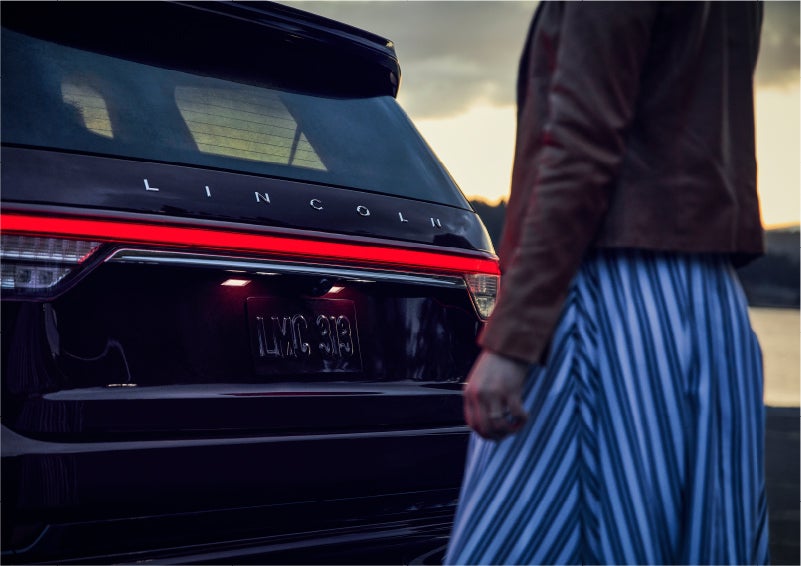 A person is shown near the rear of a 2023 Lincoln Aviator® SUV as the Lincoln Embrace illuminates the rear lights | Thornhill Lincoln in Chapmanville WV