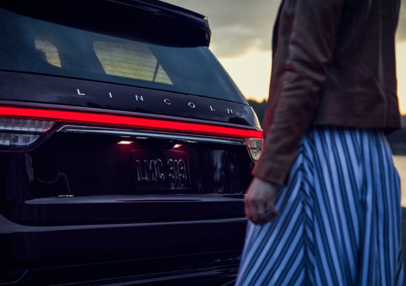 A person is shown near the rear of a 2024 Lincoln Aviator® SUV as the Lincoln Embrace illuminates the rear lights | Thornhill Lincoln in Chapmanville WV