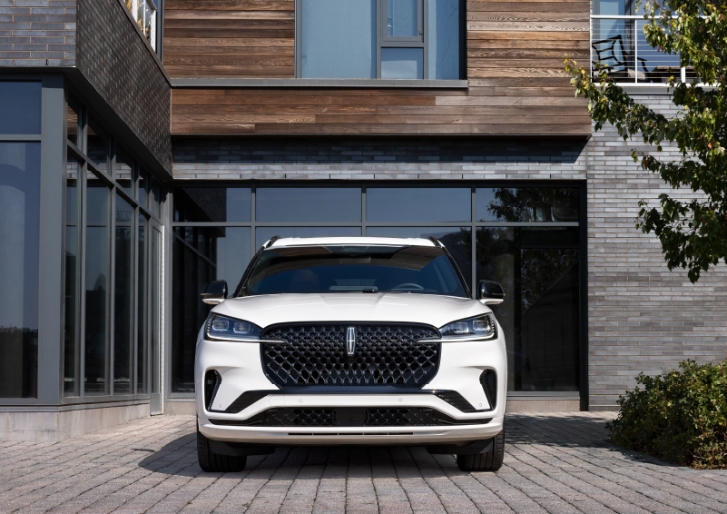 A white 2025 Lincoln Aviator® SUV is shown parked outside a modern building with gray brick and wood accents. | Thornhill Lincoln in Chapmanville WV